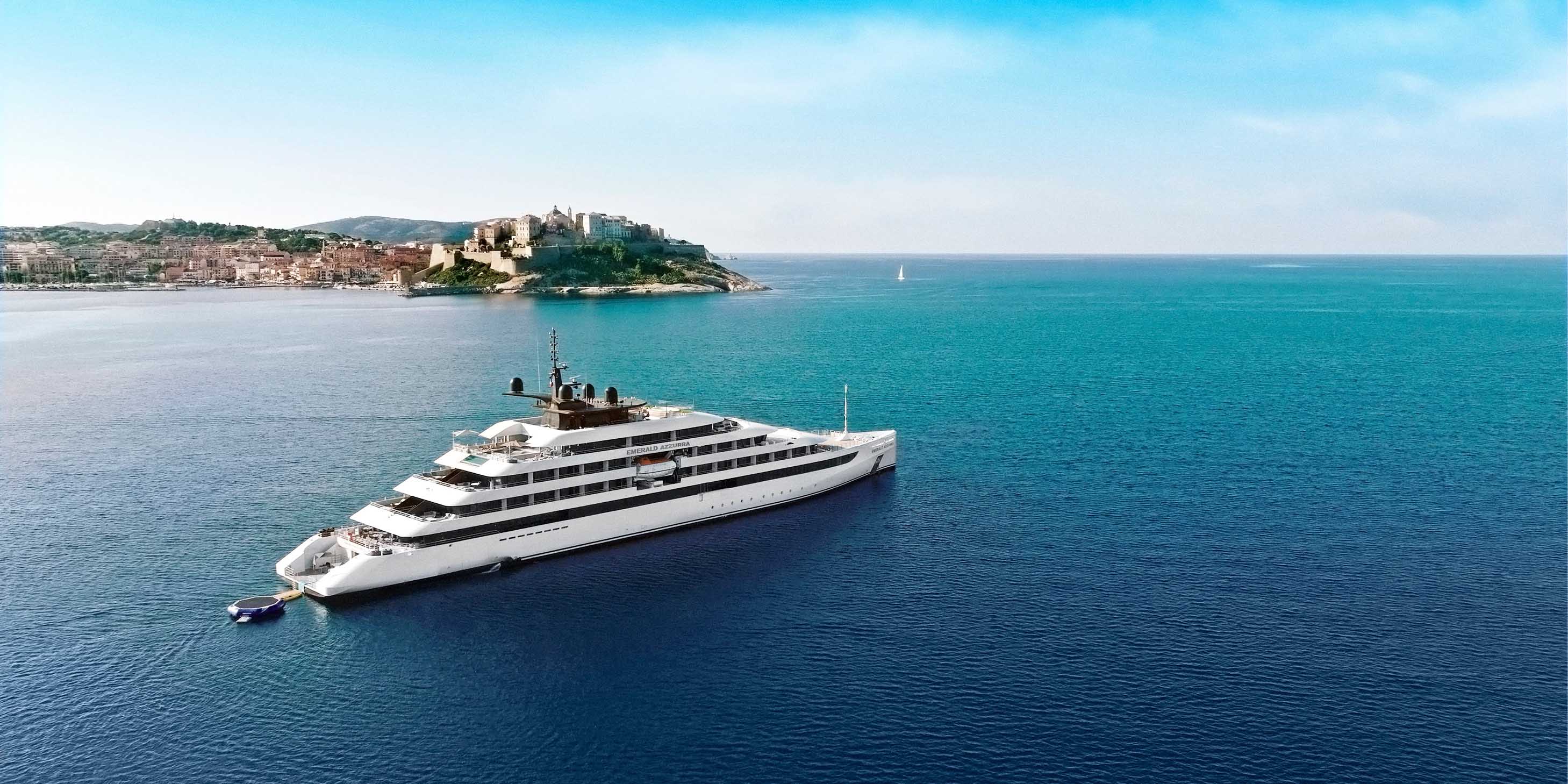 Emerald Cruises luxury yacht sailing along azure blue waters past the medieval town of Calvi nestled on the coastline on Corsica