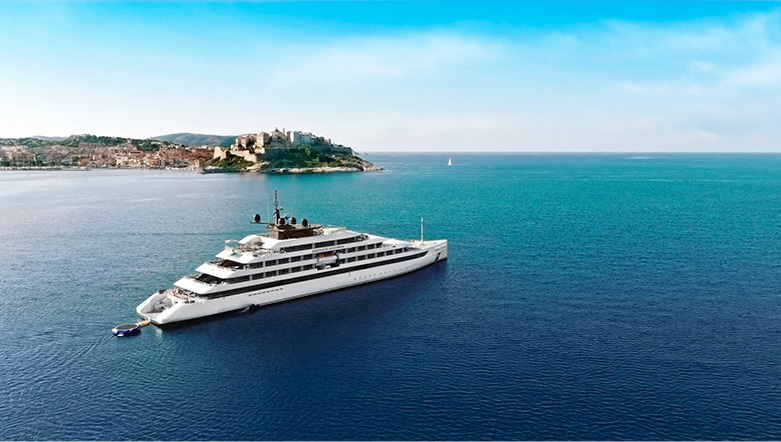 Emerald Cruises luxury yacht sailing along azure blue waters past the medieval town of Calvi nestled on the coastline on Corsica