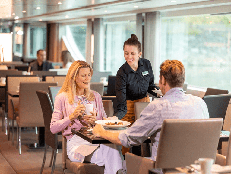 A man and woman being served plates of food by a waiter in the Crystal Dining area on board a Scenic cruise ship.