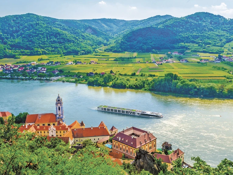 The Scenic Jasper ship cruising along the Danube River, framed by lush green fields, towering mountains, and historic buildings in Durnstein, Austria.