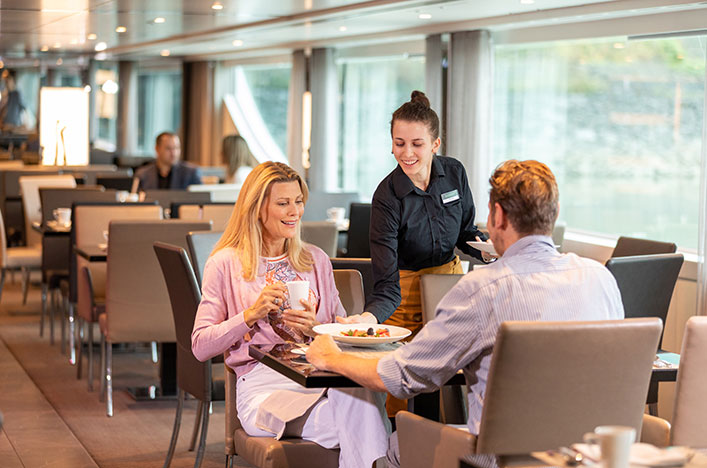 A man and woman being served plates of food by a waiter in the Crystal Dining area on board a Scenic cruise ship.  