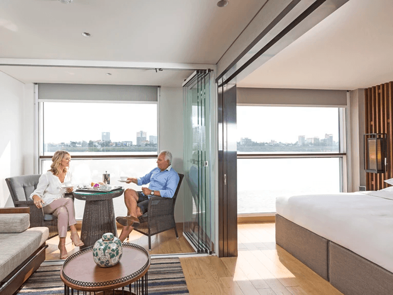  A couple relaxing in the Scenic Sun Lounge area of a Deluxe Suite, with floor-to-ceiling windows behind them with views of the river.
