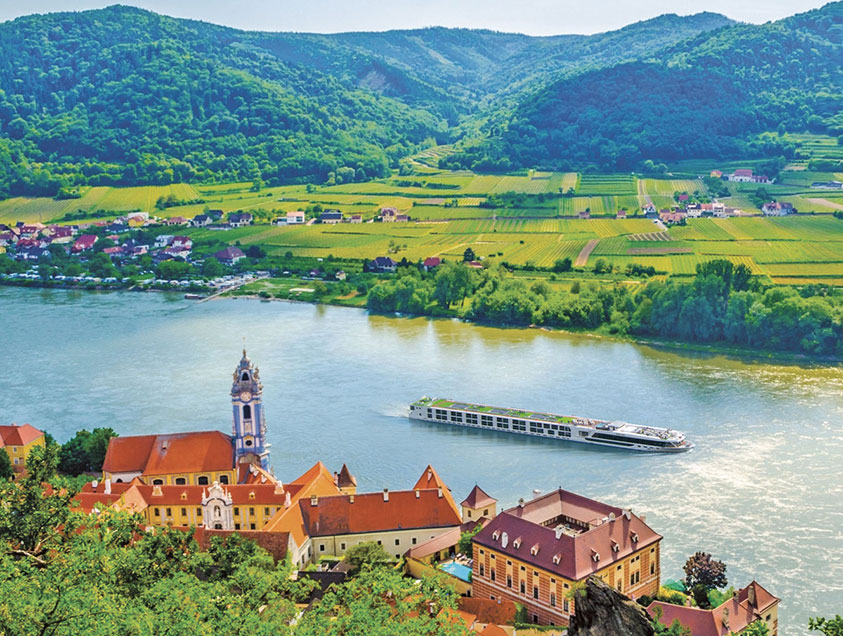 A Scenic cruise ship on the Danube River, surrounded by lush green fields and mountains, and the historical architecture of Durnstein, Austria. 