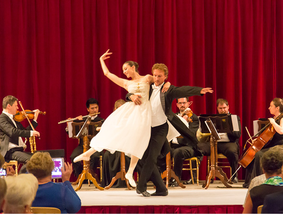 Ballet dancers perform at an exclusive concert with an orchestra and red curtains behind
